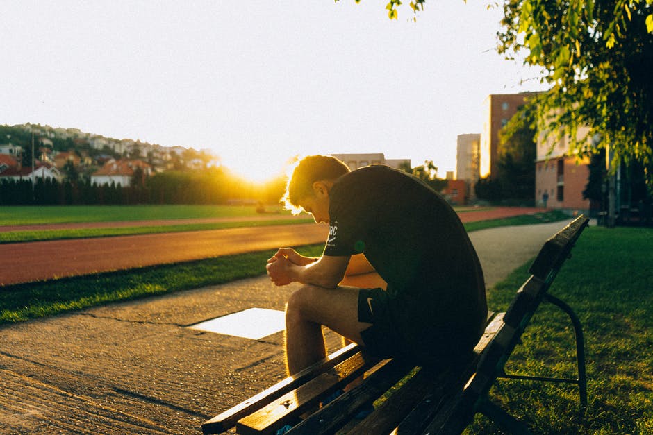 A person wearing exercise clothes, sitting on a bench, head low; there is a track field on the far plane.